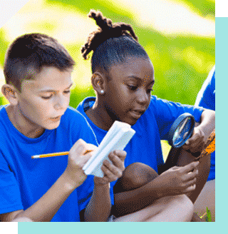 Two young people outdoors, one writing in notebook and one using magnifying glass to look at leaf