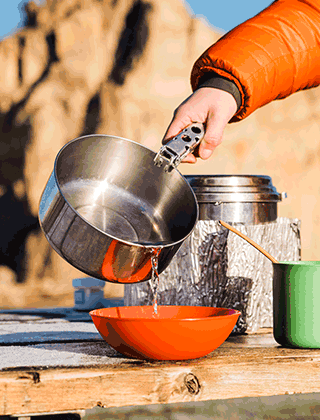 Water being poured from stainless steel pot to bowl at outdoor cooking site
