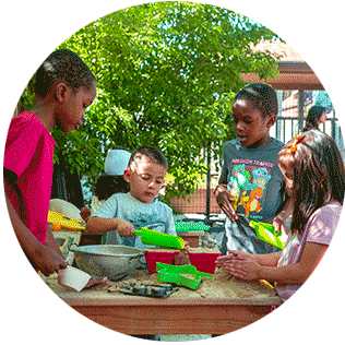 A group of four children playing with sand and baking tools on an outdoor table.