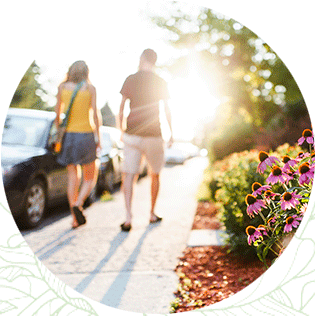 Two people pass purple coneflowers and trees while walking down a sidewalk towards a glow of sunlight.